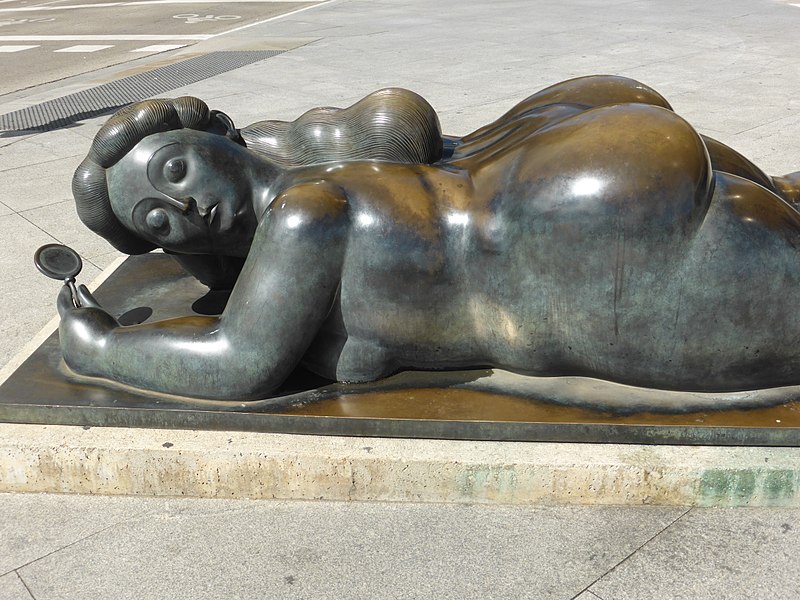 The chubby art: Fernando Botero, Woman with a mirror. Shows a sculpture of a naked chubby woman lying face down. With her face to the left holding a hand mirror in her right hand.